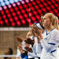 Alli stands at the end of a line of teammates during a game. They are all looking out at the court and clapping.