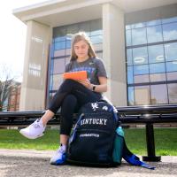 Allison sits on the bench outside the library, with her backpack on the ground beside her. She is looking at an iPad in an orange case.