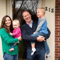 A family photo of Amberlee holding Anna, and her husband Joel holding Jacob in front of their home.