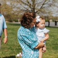 A photo of Anne Sydney holding her youngest child, Eloise, on her hip. Her husband, Jesse, can be seen in the background. Eloise is a young white girl with short blonde hair. She is wearing a white bow in her hair and a frilly white dress. Jesse is a white man with short brown hair. He is wearing a light blue short-sleeve polo, a brown belt, and khakis.