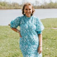 A photo of Anne Sydney smiling as she stands with one hand on her hip in front of the lake at the park.