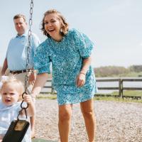 A photo of Anne Sydney and Jesse pushing their children on a swing set. Their oldest daughter, Winnie (left), is a young white girl with medium-length blonde hair. She is wearing a blue and white gingham dress that has a white collar, and a white bow in her hair.