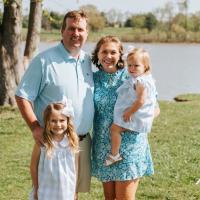 A photo of the Parrish family smiling as they pose together for the camera in front of a lake at the park.