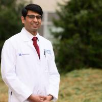 A photo of Dr. Chaitanya Iragavarapu smiling as he stands outside in front of a building on the UK HealthCare campus. He is a South Asian man with short black hair. He is wearing a white lab coat over a white button-up shirt with a red tie.