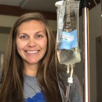 A photo of Anne Sydney smiling while posing with her IV bag and pointing her index finger in the air.