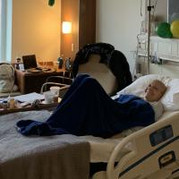 A photo of a bald Anne Sydney with a radiant smile on her face while sitting under a blanket in her hospital bed.