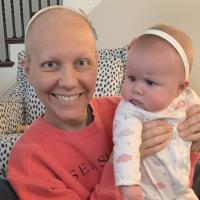 A close-up photo of a bald Anne Sydney smiling while holding up baby Eloise. They are wearing matching white headbands.