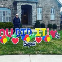 A photo of Anne Sydney smiling while standing in front of her house. There are signs in the yard that read “Congrats,” “Proud of You,” and “You Did It!”