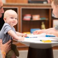 Tyler Yost holds his son Arlo while he’s coloring alongside his older brother Ezra, who is a brunette white toddler that is wearing a grey and white striped shirt.