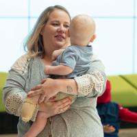 Emily Yost, a blonde woman wearing a grey shirt with white lace detailing, holds her son Arlo as he looks behind her.