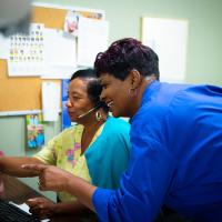 Beata leans over a nurse's shoulder while working at a computer station and points at something on screen.