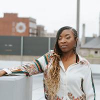 A photo of Brandi leaning against a cinder block guard rail as she looks off-camera. Buildings can be seen in the skyline behind her.