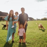 Charlie’s family—including his mom, Cassidy, wearing a teal dress, his dad, Alex, wearing shorts and a navy button-down, and his brother, Baylor, wearing shorts and a pink button-down—poses for a family photo in front of their home as their two dogs run around them.