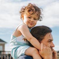 A close-up photo of Charlie sticking his tongue out while he sits on his dad’s shoulders. His dad holds onto his hands.