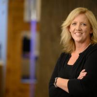 A photo of Jennifer Guilliams smiling for the camera as she poses with her arms crossed. She is a white woman with mid-length blonde hair. She is wearing a black cardigan over a black top.