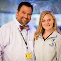 Christa and Dr. John D’Orazio pose and smile for the camera. Dr. D’Orazio is a white man with dark brown hair and scruff, he is wearing a light pink button-up shirt with a multicolor bowtie.