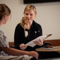 Christa sits and talks to one of her coworkers while holding some reading material. Her coworker is a white woman with brown hair in a bun, and she is wearing scrubs with a pattern on them.