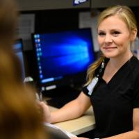 An over-the-shoulder shot of Christa smiling while sitting at a computer as she talks to her coworker.