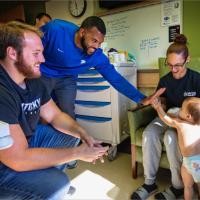 Courtney Love bends down to give a toddler a high five. There are several other football players in the room, along with the toddler’s mother.