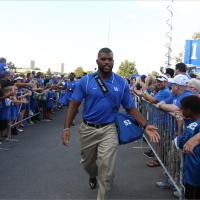 Courtney, wearing a blue polo shirt, walks down the center of two barriers with UK fans behind them. He holds out his hand to give a little boy a high five.