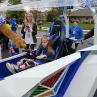 Courtney and the Wildcat mascot surround a little girl in a futuristic-looking blue and white cart. The girl has her hair in pigtails and has pink and purple leg braces.