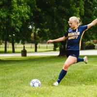 Audrey Wells, a fifteen year old blonde girl, prepares to kick a soccer ball with her arms in the air and her right leg swung behind her.