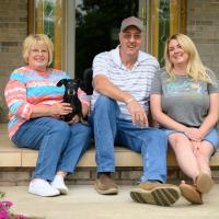 Danny, his wife, and his daughter—a middle-aged woman with blonde hair—smile and sit side by side in front of the house.