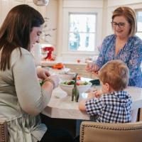 Nancy Johnson, an older white woman with blonde hair wearing a blue and pink print top, preps vegetables in her kitchen. With her is her grandson, a toddler in a blue, white and black checkered shirt, and her daughter, Kelsey, a younger white woman with brown hair, wearing a light green top.