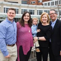 Dr. Johnson and his family smile for a picture outside UK HealthCare. Dr. Johnson, his grandson, and his son-in-law, Kyle are wearing matching blue checkered shirts, while Nancy is wearing a black top and Kelsey is wearing a plum top.