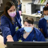 Kelsey stands beside two of her female work colleagues pointing to a computer screen. They are all wearing navy scrubs with different colored face masks.