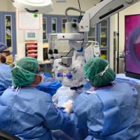 Three surgeons in an operating room: Dr. Claire Fraser, Dr. Michelle Abou-Jaoude, a middle-aged black woman, and surgical technician Jeremy Shearer, a middle-aged white man. They are performing eye surgery, wearing blue scrubs, hairnets and facemasks. They are looking at the procedure on a large screen to the right of the operating table.