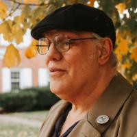 Donald stands with his face turned to the side to reveal his profile, in an outside setting under a tree with bright yellow leaves. He is wearing a black ivy hat with black-framed spectacles and a brown coat.