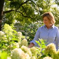 E looks at flowers in her garden. She is wearing a pinstriped button-up shirt.