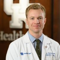 A photo of Dr. Chase Kluemper smiling for the camera while standing in front of a wall with a UK HealthCare logo. He is a young white man with short blonde hair. He is wearing a white lab coat over a blue button-up and a green tie.