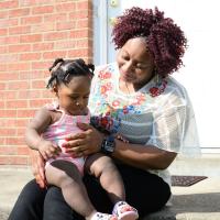 A photo of Betty sitting on her mother Deakunuh’s knee on the front step of their house. Deakunuh is a Black woman with curly red hair. She is wearing a white short-sleeve shirt with colorful embroidery on it, black pants, and gold hoop earrings.