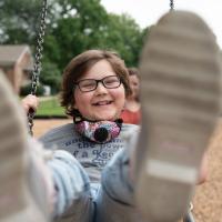 A close-up photo of Ellie swinging and smiling with her feet closest to the camera and blurred slightly. Her mother, Morgan, can be seen in the background.