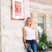 Wearing a white tank top and blue jeans, Emma stands next to a Milwaukee School of Engineering sign.