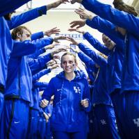 Emma, wearing a blue UK hoodie, runs between two rows of swimmers, all dressed in blue. They have their arms out, forming an arch that she’s running through.