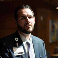 A portrait of Chase Gibony, a younger white man with dark facial hair wearing a suit jacket and tie.