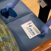 A close-up photo of a person placing a small sign onto a chair that reads ‘Do Not Use This Seat’ to enforce social distancing. They are wearing gloves and cleaning the seat.