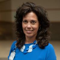 A portrait of Charlene Hale, a white woman with curly dark brown hair dressed in a blue shirt and blue jewelry, smiling at the camera.