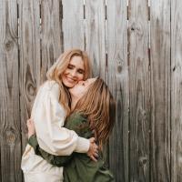 Evan embraces his mom, Krystena Richards, a middle-aged white woman with blonde hair wearing a white long-sleeve top. He gives her a peck on the cheek as she smiles broadly.