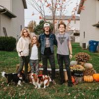 Evan standing side by side with his family and two dogs. On his right his mother, Krystena, smiles softly. On his left are his brothers, tall young adult white boys with medium to long brown hair. One is wearing a white baseball cap backwards and a denim jacket with black jeans. The other is wearing a grey shirt with black jeans. All are smiling.