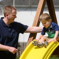 A picture of Everett sitting at the top of a slide, Daniel is standing next to him and has his arm on Everett’s back.