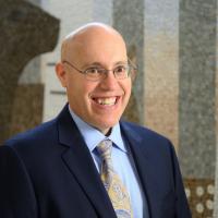 A photo of Dr. Hubie Ballard enthusiastically smiling while standing in front of a mosaic. He is a white bald man who is wearing glasses, a navy suit jacket, a light blue shirt, and a paisley tie.