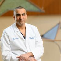 Dr. Malay Shah looks into the camera. He is a man of Middle Eastern descent with graying short black hair, he is wearing a white lab coat over blue scrubs.