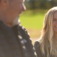 A shot of DeeAnn smiling as she stands outside, Fred is out of focus in the foreground.