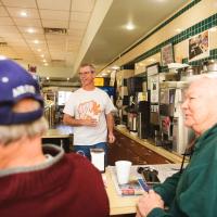 Fred walks behind the serving counter over towards some patrons sitting at the counter.