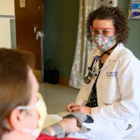 Geri sits across from a patient in a hospital room, wearing her facemask and goggles. It’s clear that she’s smiling under her mask.