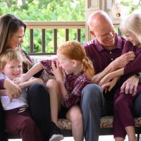 A candid photo of A.W. and his family as they play. His wife is a white woman with brunette hair, who is wearing a burgundy sweater. His son is a young white child with blonde hair, he is wearing a white shirt and burgundy pants. His daughter sitting in the middle is a young white girl with orange hair in pigtails, she is wearing a burgundy plaid shirt. His daughter that is sitting on his knee is a young white girl with light blonde hair, she is wearing a burgundy romper.